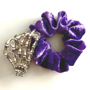 Puede incluir: Una pulsera plateada con forma de mano de esqueleto junto a una goma para el pelo de terciopelo morado. La goma para el pelo es de un tono morado intenso y tiene un aspecto suave y texturizado. La pulsera est&aacute; detallada con huesos de dedos individuales y una calavera en la mu&ntilde;eca.
