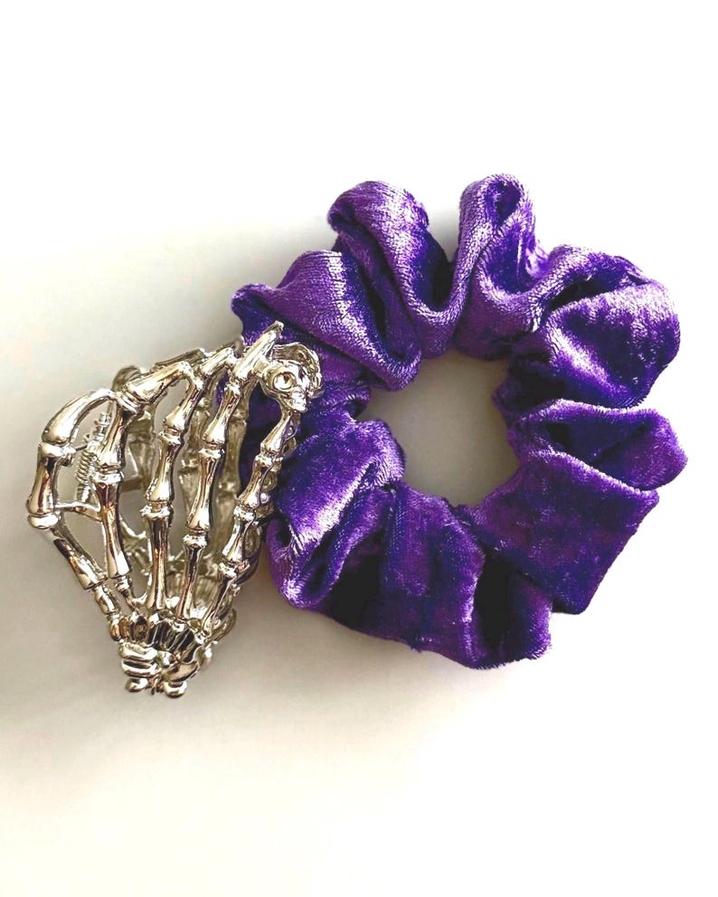 Puede incluir: Una pulsera plateada con forma de mano de esqueleto junto a una goma para el pelo de terciopelo morado. La goma para el pelo es de un tono morado intenso y tiene un aspecto suave y texturizado. La pulsera est&aacute; detallada con huesos de dedos individuales y una calavera en la mu&ntilde;eca.