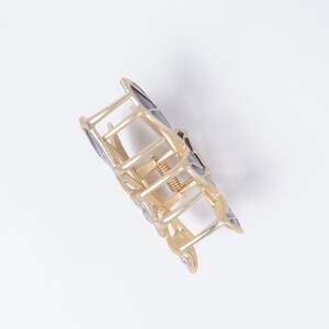 May include: Gold-toned metal hair claw clip with geometric black and gray accents. The clip features a spring mechanism and a series of interlocking bars forming a decorative shape. This hair accessory is designed to hold hair in place.