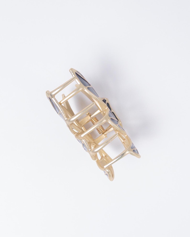 May include: Gold-toned metal hair claw clip with geometric black and gray accents. The clip features a spring mechanism and a series of interlocking bars forming a decorative shape. This hair accessory is designed to hold hair in place.