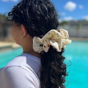 May include: A cream-colored scrunchie with a floral pattern is worn in a ponytail. The scrunchie is made of a soft fabric and features small brown and green floral designs. The person is wearing a white shirt, and the background shows a pool and blue sky.