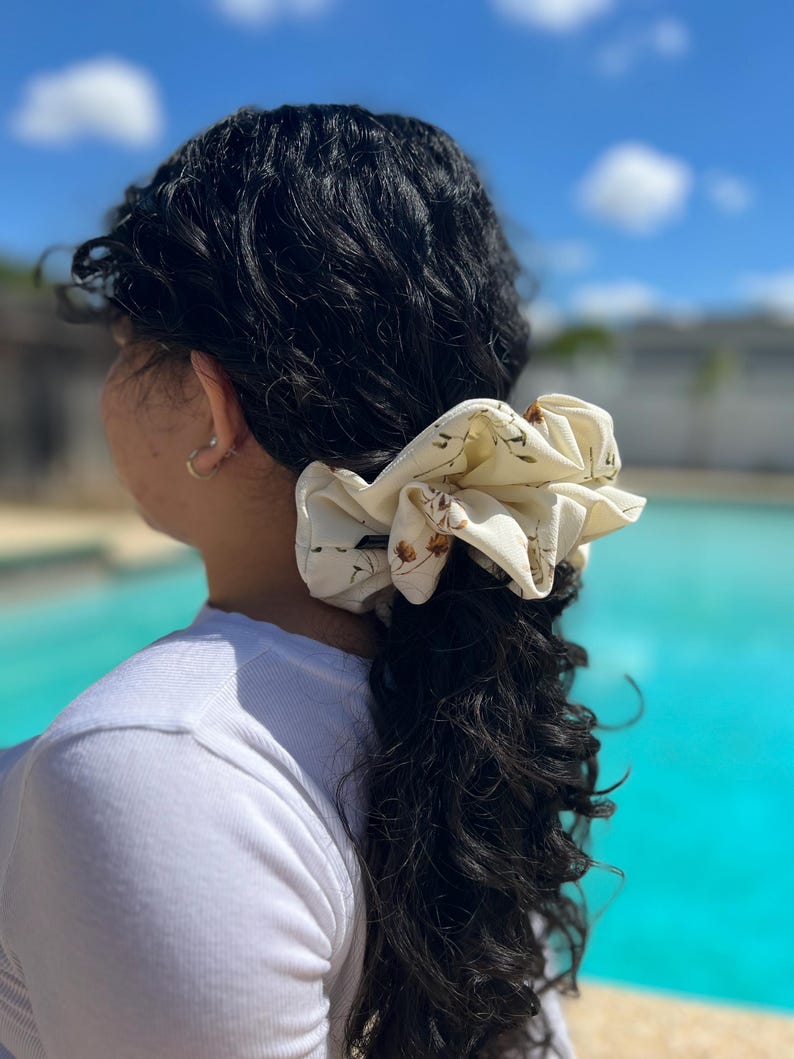 May include: A cream-colored scrunchie with a floral pattern is worn in a ponytail. The scrunchie is made of a soft fabric and features small brown and green floral designs. The person is wearing a white shirt, and the background shows a pool and blue sky.