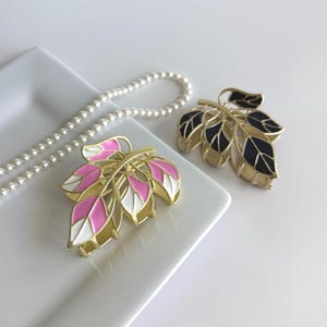 May include: Two leaf-shaped hair clips with gold-tone metal frames. One clip features pink and white enamel leaves, while the other has black enamel leaves. A pearl necklace is in the background.