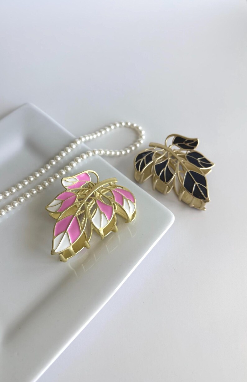May include: Two leaf-shaped hair clips with gold-tone metal frames. One clip features pink and white enamel leaves, while the other has black enamel leaves. A pearl necklace is in the background.