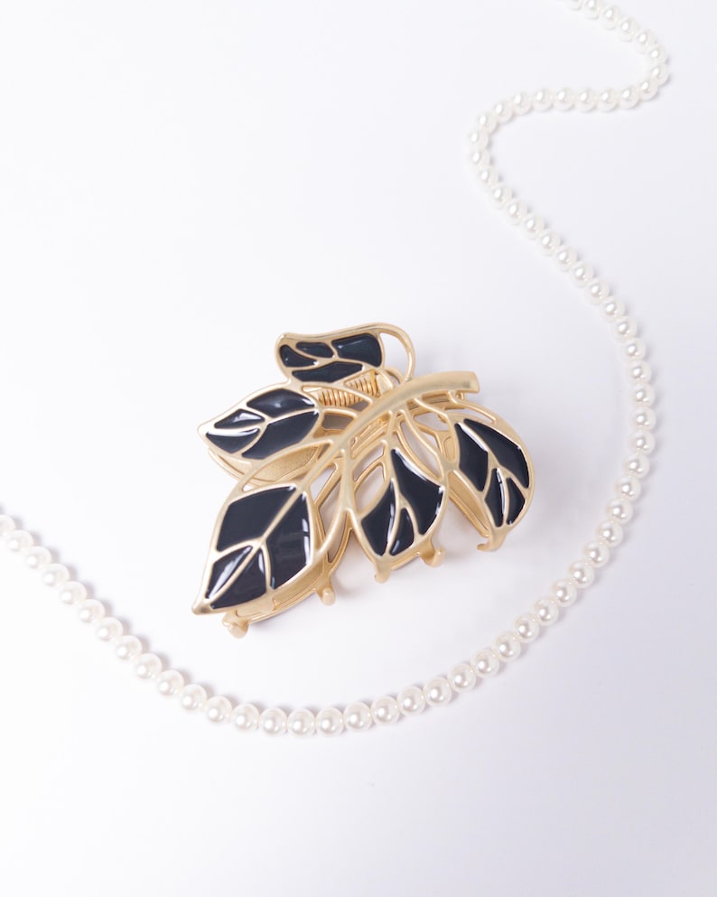 May include: A gold-toned metal hair claw clip in a leaf design with black enamel detailing. A strand of white faux pearls curves across the white background, adding a touch of elegance to the hair accessory.