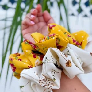 May include: A collection of fabric scrunchies in various floral patterns. One scrunchie is yellow with white polka dots and red flowers. Another is white with brown floral designs. The scrunchies are held in a hand.