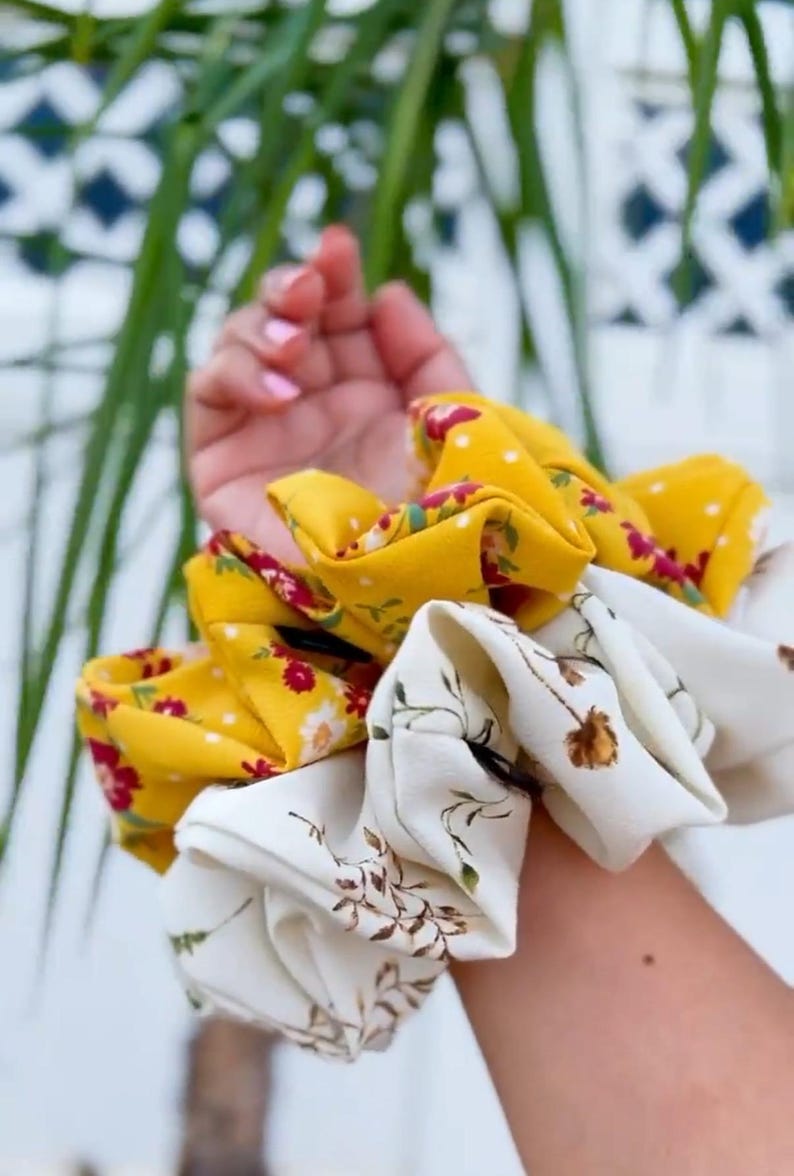 May include: A collection of fabric scrunchies in various floral patterns. One scrunchie is yellow with white polka dots and red flowers. Another is white with brown floral designs. The scrunchies are held in a hand.