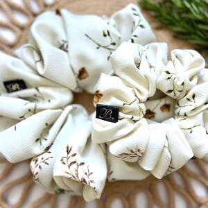 May include: Two ivory scrunchies with a floral pattern of brown and green leaves. Each scrunchie has a black label with a white initial. The scrunchies are arranged on a woven, tan surface.
