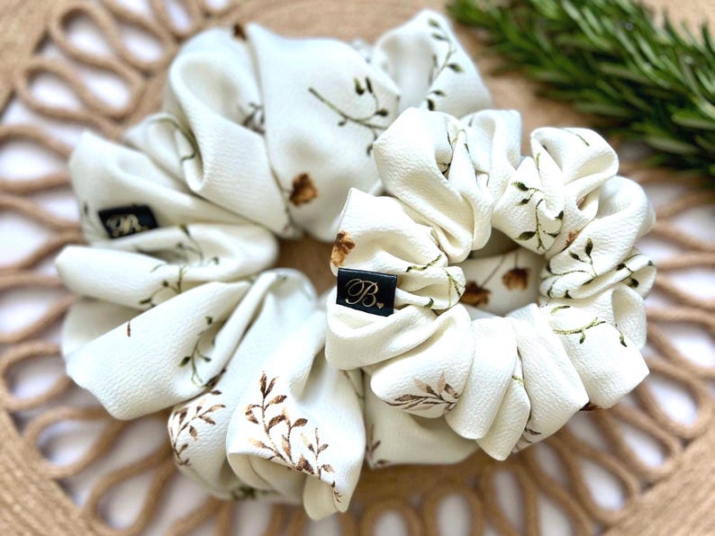 May include: Two ivory scrunchies with a floral pattern of brown and green leaves. Each scrunchie has a black label with a white initial. The scrunchies are arranged on a woven, tan surface.