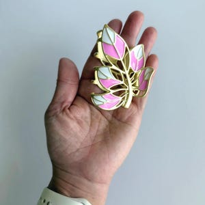 May include: A gold-colored leaf-shaped hair clip with pink and white enamel detailing. The clip is held in a hand, showcasing its design and color scheme. The clip's design is reminiscent of a stylized leaf.