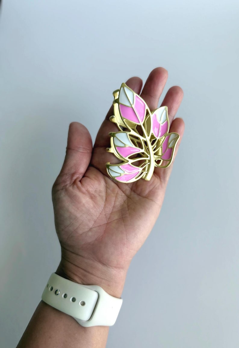 May include: A gold-colored leaf-shaped hair clip with pink and white enamel detailing. The clip is held in a hand, showcasing its design and color scheme. The clip's design is reminiscent of a stylized leaf.