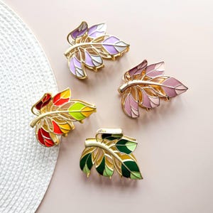 May include: Four leaf-shaped hair clips in various colors. Each clip features a gold-colored metal frame with enamel-filled leaves in shades of purple, pink, red, yellow, green, and white. The clips are arranged on a light pink surface.