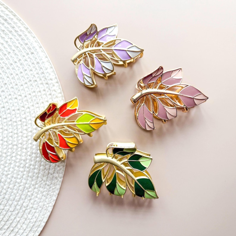 May include: Four leaf-shaped hair clips in various colors. Each clip features a gold-colored metal frame with enamel-filled leaves in shades of purple, pink, red, yellow, green, and white. The clips are arranged on a light pink surface.