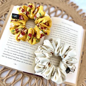 May include: Two scrunchies, one yellow with red floral print and one cream with brown floral print, rest on an open book. The yellow scrunchy has a black tag with a white "B" on it. The book is open to page 46.