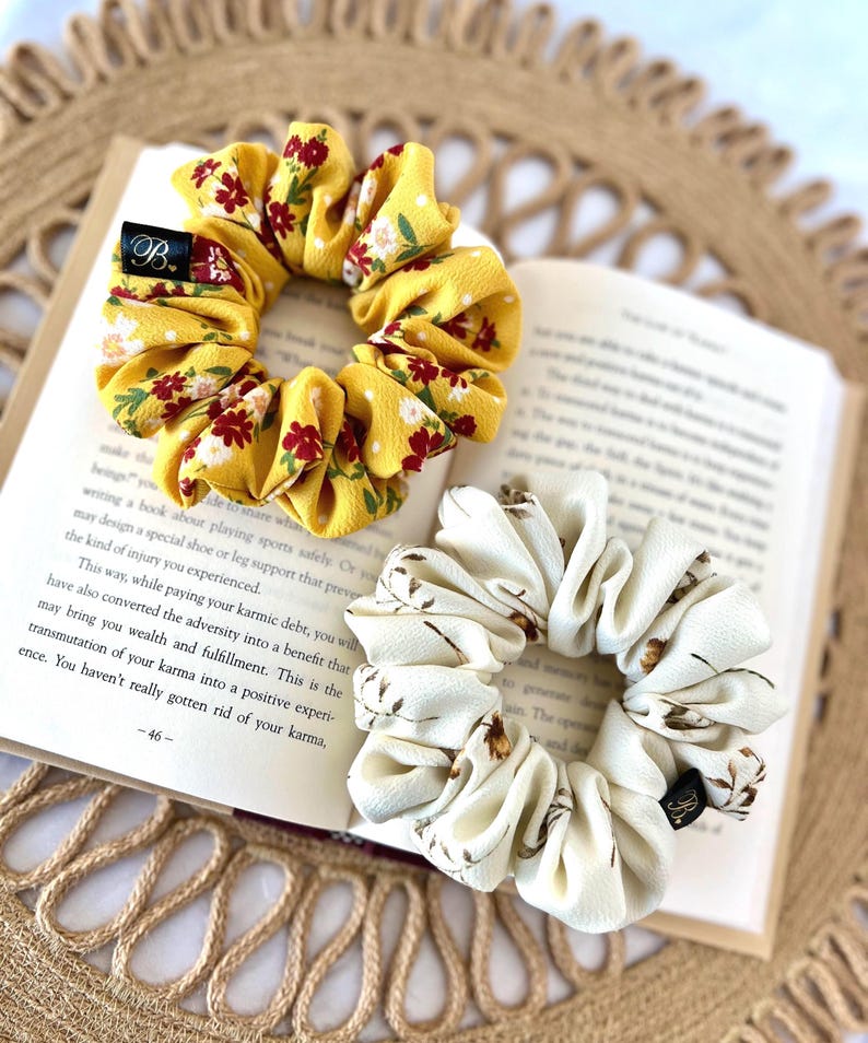 May include: Two scrunchies, one yellow with red floral print and one cream with brown floral print, rest on an open book. The yellow scrunchy has a black tag with a white "B" on it. The book is open to page 46.
