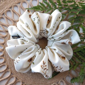 May include: A large, ivory scrunchie with a floral pattern in brown and green. The fabric is gathered around a neutral-colored elastic band. A small black tag with white text is visible on the side. The scrunchie is a hair accessory.
