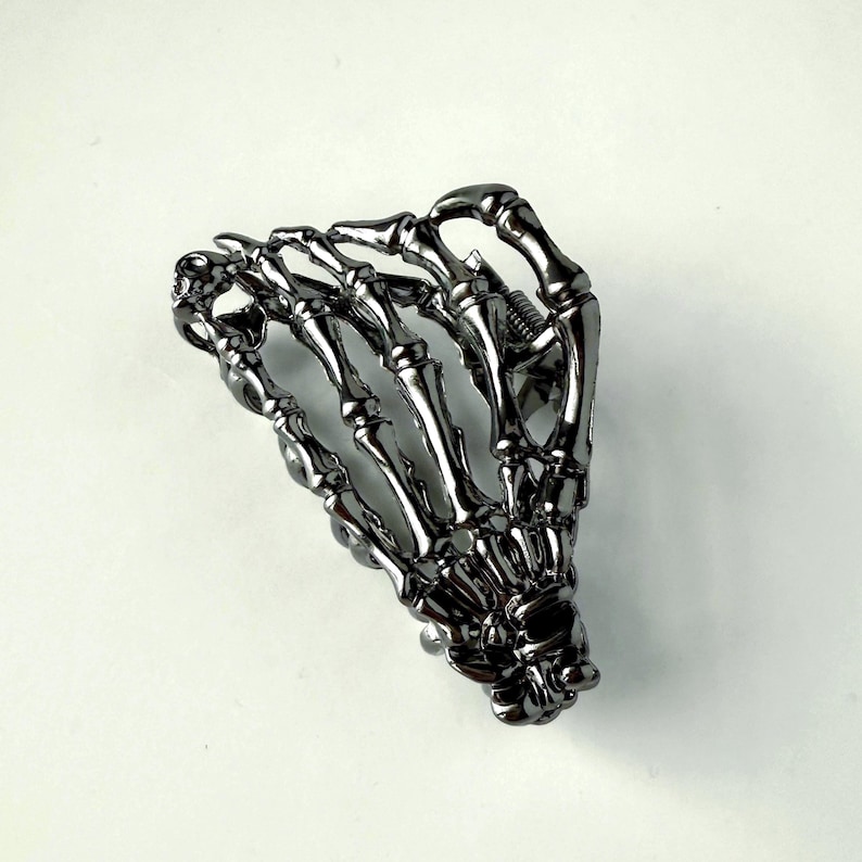 May include: A silver skeleton hand hair clip. The clip is made of metal and is shaped like a hand with the bones showing. It is a decorative hair accessory.