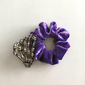 Puede incluir: Una coletero de terciopelo morado junto a un accesorio para el cabello de mano de esqueleto plateada. El coletero es de un tono morado vibrante y tiene un aspecto suave y texturizado. La mano de esqueleto a&ntilde;ade un toque g&oacute;tico.