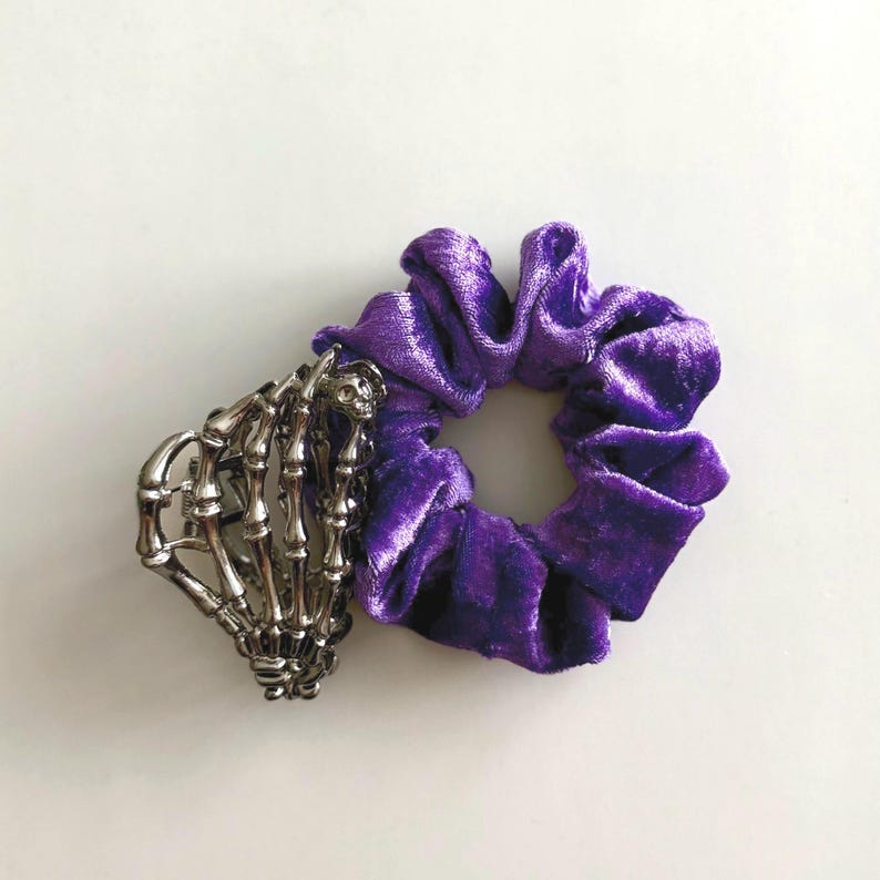 Puede incluir: Una coletero de terciopelo morado junto a un accesorio para el cabello de mano de esqueleto plateada. El coletero es de un tono morado vibrante y tiene un aspecto suave y texturizado. La mano de esqueleto a&ntilde;ade un toque g&oacute;tico.