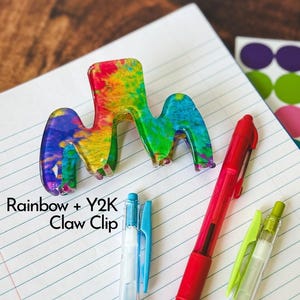 May include: A rainbow-colored claw clip with the text "Rainbow + Y2K Claw Clip" is displayed on a lined notebook paper. The clip is next to a red pen, a blue pen, and a green pen.