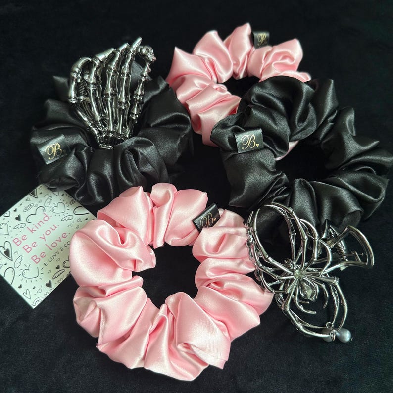 Puede incluir: Colecci&oacute;n de coleteros de sat&eacute;n en negro y rosa, con accesorios met&aacute;licos decorativos para el cabello. Un coletero presenta una mano de esqueleto, otro una ara&ntilde;a, y otros tienen una peque&ntilde;a etiqueta con la letra "B". Tambi&eacute;n hay una tarjeta con el texto "Be kind. Be you. Be love."