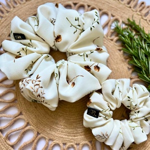 May include: Two ivory scrunchies with a delicate floral pattern in brown and green. The scrunchies are made of a soft fabric and are arranged on a woven mat. One scrunchie is larger than the other.
