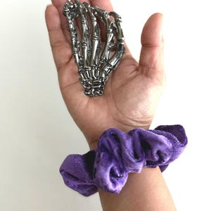 Puede incluir: Una mano de esqueleto plateada descansa en una palma abierta, con una scrunchie de terciopelo morado en la mu&ntilde;eca. La mano es detallada. La scrunchie es de un morado intenso.