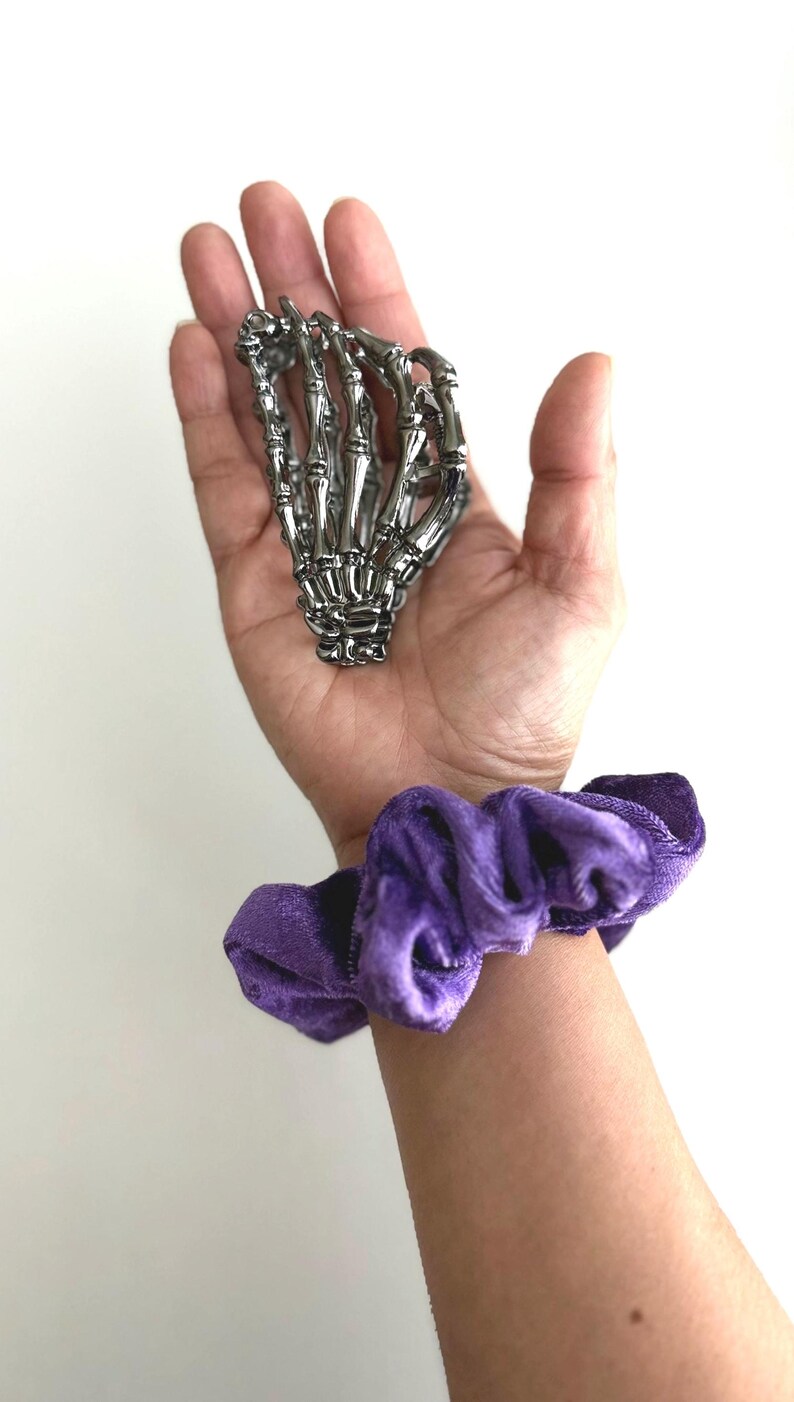 Puede incluir: Una mano de esqueleto plateada descansa en una palma abierta, con una scrunchie de terciopelo morado en la mu&ntilde;eca. La mano es detallada. La scrunchie es de un morado intenso.