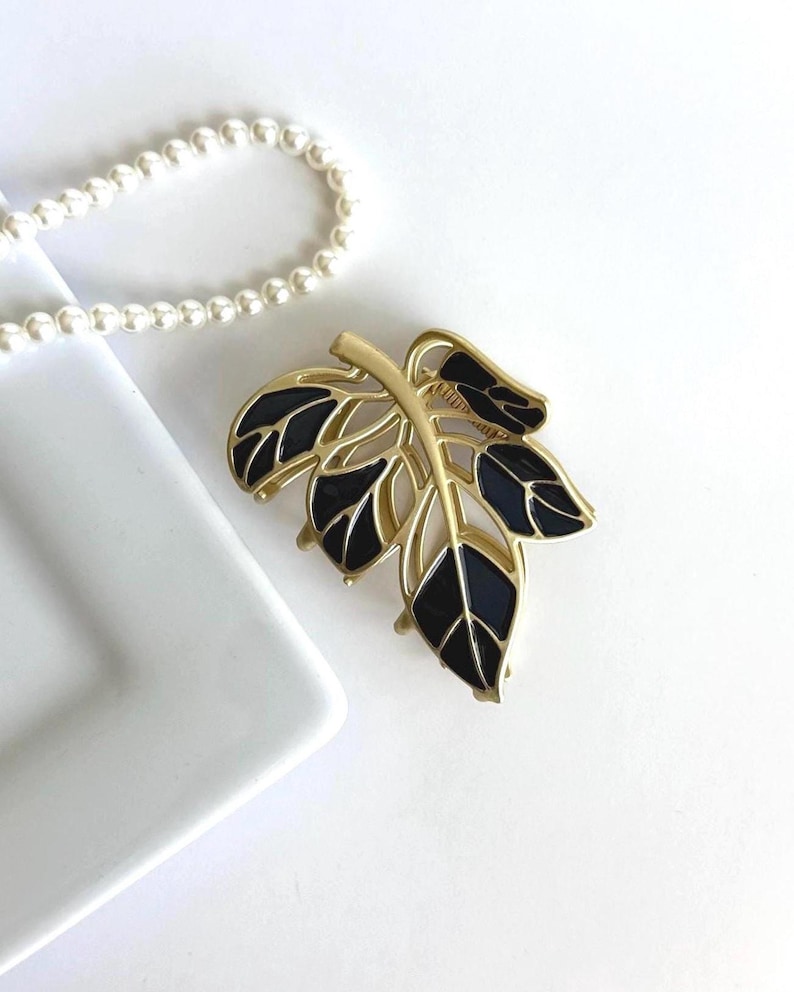 May include: Gold-toned leaf brooch with black enamel detailing. The brooch features a detailed leaf design with a gold stem and veins. A white pearl necklace is in the background. Jewelry.