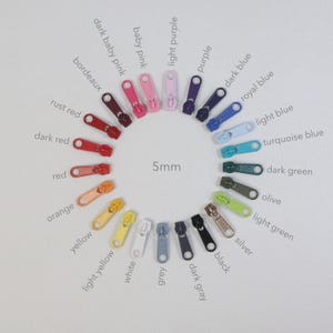 Könnte beinhalten: Eine Sammlung von 5mm Reißverschlüssen in verschiedenen Farben, kreisförmig angeordnet. Zu den Farben gehören Rot, Orange, Gelb, Grün, Blau, Lila und neutrale Töne wie Schwarz, Weiß und Grau. Jeder Reißverschluss ist mit seinem Farbnamen beschriftet.