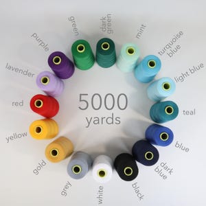 Puede incluir: Una disposición circular de diecisiete carretes de hilo en varios colores, incluyendo morado, verde, menta, turquesa, azul, amarillo, rojo, dorado, gris, blanco y negro. El texto 5000 yards está impreso en el centro de la rueda de color.