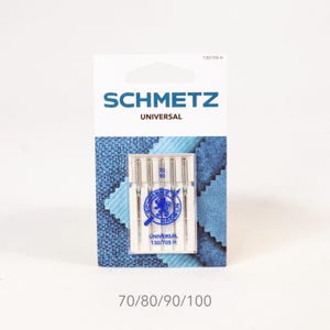 Könnte beinhalten: Eine Packung SCHMETZ UNIVERSAL Nähmaschinennadeln. Die weiß-blaue Verpackung enthält fünf Nadeln, mit den Nadelgrößen 70/80/90/100 unten aufgedruckt. Die Packung trägt auch den Text "UNIVERSAL 130/705 H".