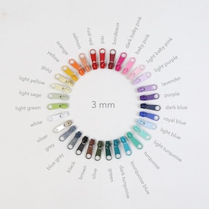 May include: A selection of 3 mm zippers in a circular pattern, showcasing a spectrum of colours like red, green, and blue. The image features zippers in shades such as "orange," "lavender," and "turquoise." The zippers are displayed against a white backdrop.