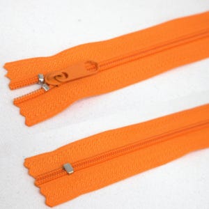 Può includere: Primo piano di due cerniere arancioni. Una cerniera è aperta, rivelando i denti, mentre l'altra è chiusa. I tiranti sono arancioni e i cursori metallici sono argentati.