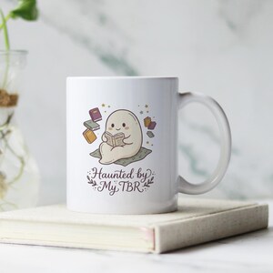 Op de afbeelding: Een witte keramische mok met een cartoonillustratie van een kleine, ronde, beige geest die op een kussen zit en een boek leest. Verschillende kleurrijke boeken zweven rond de geest. De tekst "Haunted by My TBR" is in donkerbruin gedrukt.