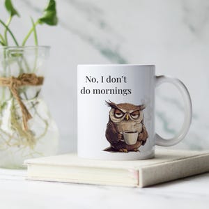 Op de afbeelding: Een witte keramische koffiemok met een illustratie van een bruine uil die een kopje koffie vasthoudt. Op de mok staat de tekst "No, I don't do mornings".