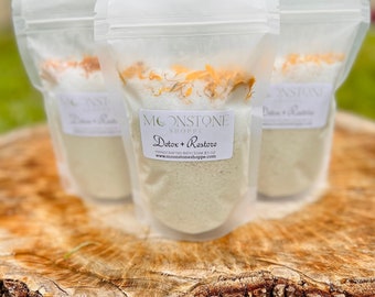May include: Three clear plastic bags filled with white bath salts and orange flower petals. The bags are labeled "Moonstone Shoppe Detox + Restore Handmade Bath Soak 8.5 oz www.moonstoneshoppe.com"