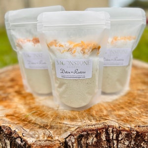 May include: Three clear plastic bags filled with white bath salts and orange flower petals. The bags are labeled "Moonstone Shoppe Detox + Restore Handmade Bath Soak 8.5 oz www.moonstoneshoppe.com"