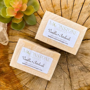 Puede incluir: Dos barras de jabón artesanal con etiquetas que dicen "Vanilla + Patchouli" y "MOONSTONE SHOPPE". Cada jabón pesa aproximadamente 99 g. El fondo es una superficie de madera con una suculenta y un cristal transparente.