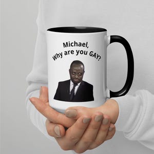 Può includere: Tazza in ceramica bianca con interno e manico neri. La tazza presenta il testo "Michael, Why are you GAY?" insieme all'immagine di un uomo in giacca e cravatta. La tazza è tenuta da una persona.