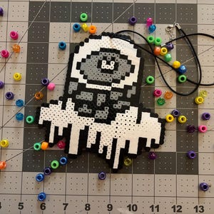 May include: A pixelated pendant necklace in black, white, and gray, featuring a unique design. The necklace has a black cord and is adorned with colorful beads. Scattered around are various colorful beads.