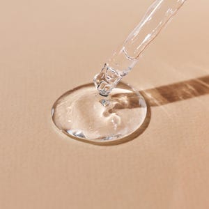 May include: A clear glass dropper dispensing a transparent liquid onto a beige surface, forming a large droplet. The dropper is angled, with the liquid creating a circular shape. The image is well-lit, highlighting the transparency of the liquid.