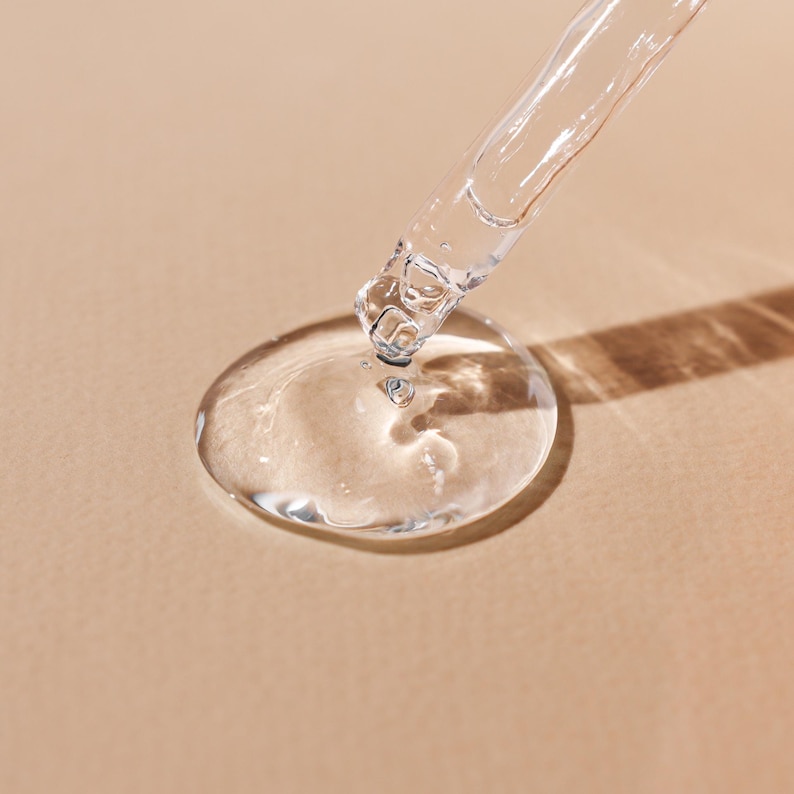 May include: A clear glass dropper dispensing a transparent liquid onto a beige surface, forming a large droplet. The dropper is angled, with the liquid creating a circular shape. The image is well-lit, highlighting the transparency of the liquid.