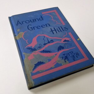 May include: A blue book with the title "Around Green Hills" printed in white. The cover features a pink and blue illustration of rolling hills with a farm scene.