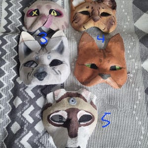 Könnte beinhalten: Fünf handgefertigte Katzenmasken in verschiedenen Farben und Designs. Die Masken bestehen aus einem strukturierten Material und weisen bemalte Details wie Augen, Nasen und Fellmuster auf. Die Masken werden auf einem gemusterten Stoffhintergrund präsentiert.