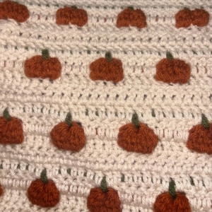 Puede incluir: Una tela de ganchillo de color crema con un patrón repetitivo de pequeñas calabazas naranjas texturizadas con tallos de color verde oscuro. Las calabazas están dispuestas en filas horizontales sobre el fondo de hilo blanco roto.