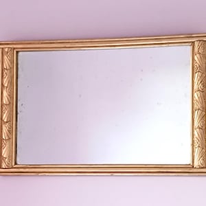 Peut inclure: Miroir rectangulaire avec un cadre doré orné de motifs sur les côtés. Le miroir est fixé sur un mur violet clair. Le cadre présente un design classique, ajoutant une touche d'élégance à la décoration.