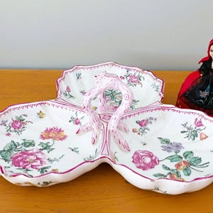 Może przedstawiać: Biała porcelanowa miska do serwowania z trzema sekcjami i różowym wzorem kwiatowym. Miska ma różowy uchwyt w środku i jest ozdobiona różowymi i zielonymi wzorami kwiatowymi.