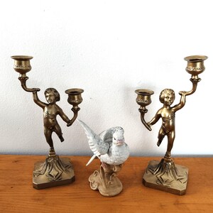 Könnte beinhalten: Zwei antike Messing-Kerzenhalter mit Cherubfiguren, die Kerzenschalen halten. Zwischen den Kerzenhaltern befindet sich eine weiße Keramik-Taubenfigur. Die Kerzenhalter haben einen quadratischen Sockel und stehen auf einer Holzoberfläche.