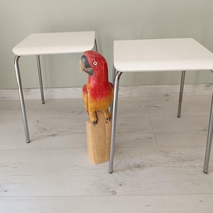 May include: Two white square-top side tables with chrome legs. A carved wooden parrot statue in red, yellow, and orange stands between the tables. The tables are suitable for a living room or bedroom.